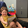 toddler, child, smile, yellow_outfit, book, dinosaurs, toy, carpet, indoor, person, happy, play, reading, feet, curly_hair, appliance, wall, floor, sitting, lap