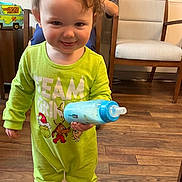 Aiden is registered to the contest to win money with this photo: toddler, child, baby_bottle, green_pajamas, smile, indoor, wooden_floor, chair, person, face, curly_hair, room, furniture, lunchbox, toy, happy, clothing, standing, cute, playful