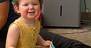 Aiden is registered to the contest to win money with this photo: appliance, book, carpet, casual, child, curly_hair, electrical_outlet, floor, green_toy, happy, indoor, person, play, reading, sitting, smiling, toddler, toy, wall, yellow_clothing