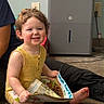 Aiden is registered to the contest to win money with this photo: toddler, child, curly_hair, yellow_clothing, carpet, book, toy, green_toy, indoor, appliance, wall, electrical_outlet, smiling, sitting, person, floor, reading, happy, play, casual