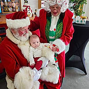 Brody is registered to the contest to win money with this photo: baby, child, santa_claus, mrs_claus, christmas, holiday, festive_clothing, red_coat, white_beard, glasses, indoor, decorations, snowman, candy_cane, plant, table, smiling, holding, person, celebration