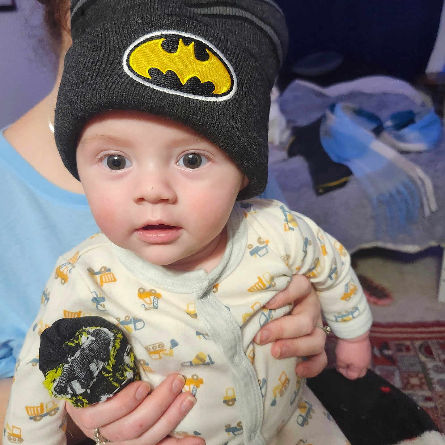 Brody is registered to the contest to win money with this photo: adult_hand, baby, background, batman_logo, beanie, child, clothing, construction_vehicle_pattern, cozy, curious, face, hand, hat, holding, indoor, onesie, person, portrait, soft_lighting, wide_eyes