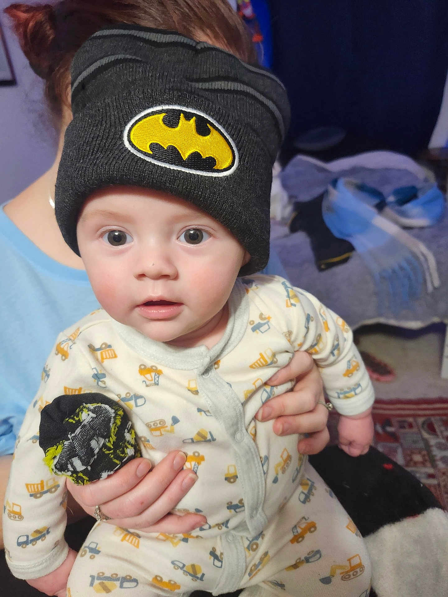 Brody is registered to the contest to win money with this photo: baby, child, beanie, hat, batman_logo, onesie, construction_vehicle_pattern, indoor, person, hand, face, wide_eyes, curious, soft_lighting, adult_hand, holding, cozy, background, clothing, portrait