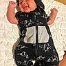 baby, infant, sleeping, peaceful, smiling, portrait, child, onesie, hood, black_clothing, origami_pattern, zipper, quilt, blanket, bed, lying_down, raised_fists, bare_feet, indoor, cozy