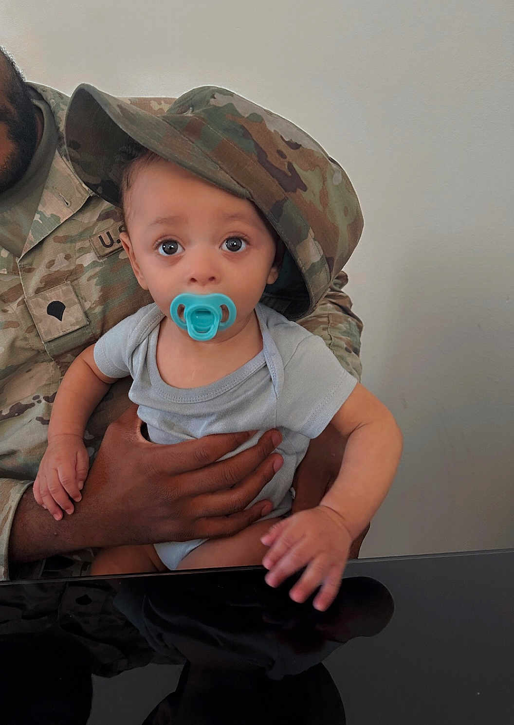 Azaylen is registered to the contest to win money with this photo: baby, pacifier, hat, camouflage, military_uniform, adult_hand, infant, child, cute, holding, indoor, face, expression, person, clothing, table, skin, eyes, portrait, casual