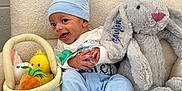 Azaylen is registered to the contest to win money with this photo: baby, child, blue_hat, bunny_ears, plush_toy, stuffed_animal, easter_basket, basket, toy_carrot, toy_duck, gray_bunny, soft_toy, cozy_chair, striped_blanket, happy, smiling, indoors, cute, infant, playful