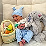baby, child, blue_hat, bunny_ears, plush_toy, stuffed_animal, easter_basket, basket, toy_carrot, toy_duck, gray_bunny, soft_toy, cozy_chair, striped_blanket, happy, smiling, indoors, cute, infant, playful