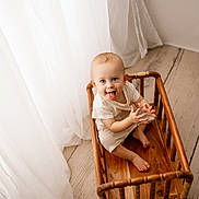 Kingston is registered to the contest to win money with this photo: baby, child, wooden_cradle, tongue_out, playful, curtains, natural_light, flooring, indoor, smiling, barefoot, cute, infant, wood, home, light, portrait, sitting, white_clothing, young_child