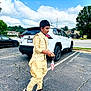 boy, child, traditional_clothing, parking_lot, car, suv, cloudy_sky, sidewalk, asphalt, sneakers, holding_object, candy, tree, outdoor, daytime, person, walking, casual, vehicle, black_shoes