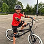 Aayan is registered to the contest to win money with this photo: child, bicycle, helmet, protective_gear, elbow_pads, knee_pads, red_shirt, black_shorts, black_bike, white_tires, parking_lot, asphalt, outdoor, trees, overcast_sky, smiling, sport, active, person, footwear