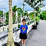 child, boy, palm_tree, water_fountain, plants, outdoor, potted_plant, sidewalk, street, cloudy_sky, greenery, casual_clothing, blue_shirt, black_shorts, sneakers, hands_in_pockets, smiling, young, daytime, nature