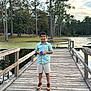 Aayan is registered to the contest to win money with this photo: boy, child, pier, lake, water, trees, outdoor, nature, wood, casual_clothing, shorts, shirt, sneakers, smile, daylight, sky, clouds, forest, person, standing