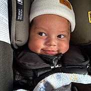 Jaxon is registered to the contest to win money with this photo: baby, child, car_seat, hat, white_hat, blanket, smile, face, infant, closeup, cute, warm_clothing, straps, safety, indoors, expression, cheeky, young_child, cozy, seat