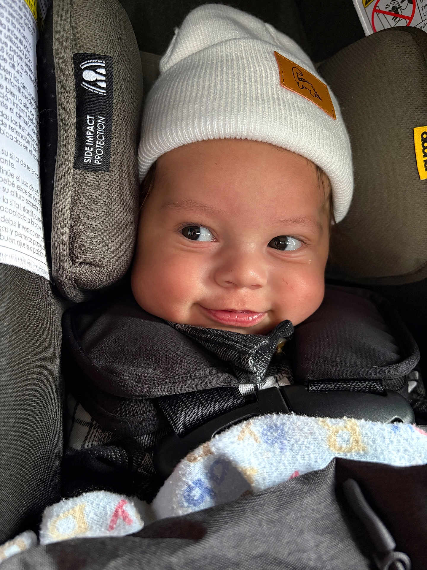 Jaxon is registered to the contest to win money with this photo: baby, child, car_seat, hat, white_hat, blanket, smile, face, infant, closeup, cute, warm_clothing, straps, safety, indoors, expression, cheeky, young_child, cozy, seat