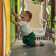 Alessio participe au concours pour gagner de l'argent avec cette photo : toddler, child, playground, smiling, kneeling, green, white_shirt, green_pants, outdoor, play_equipment, metal, wood, handrail, tree, happy, person, sock, platform, crawling, daylight