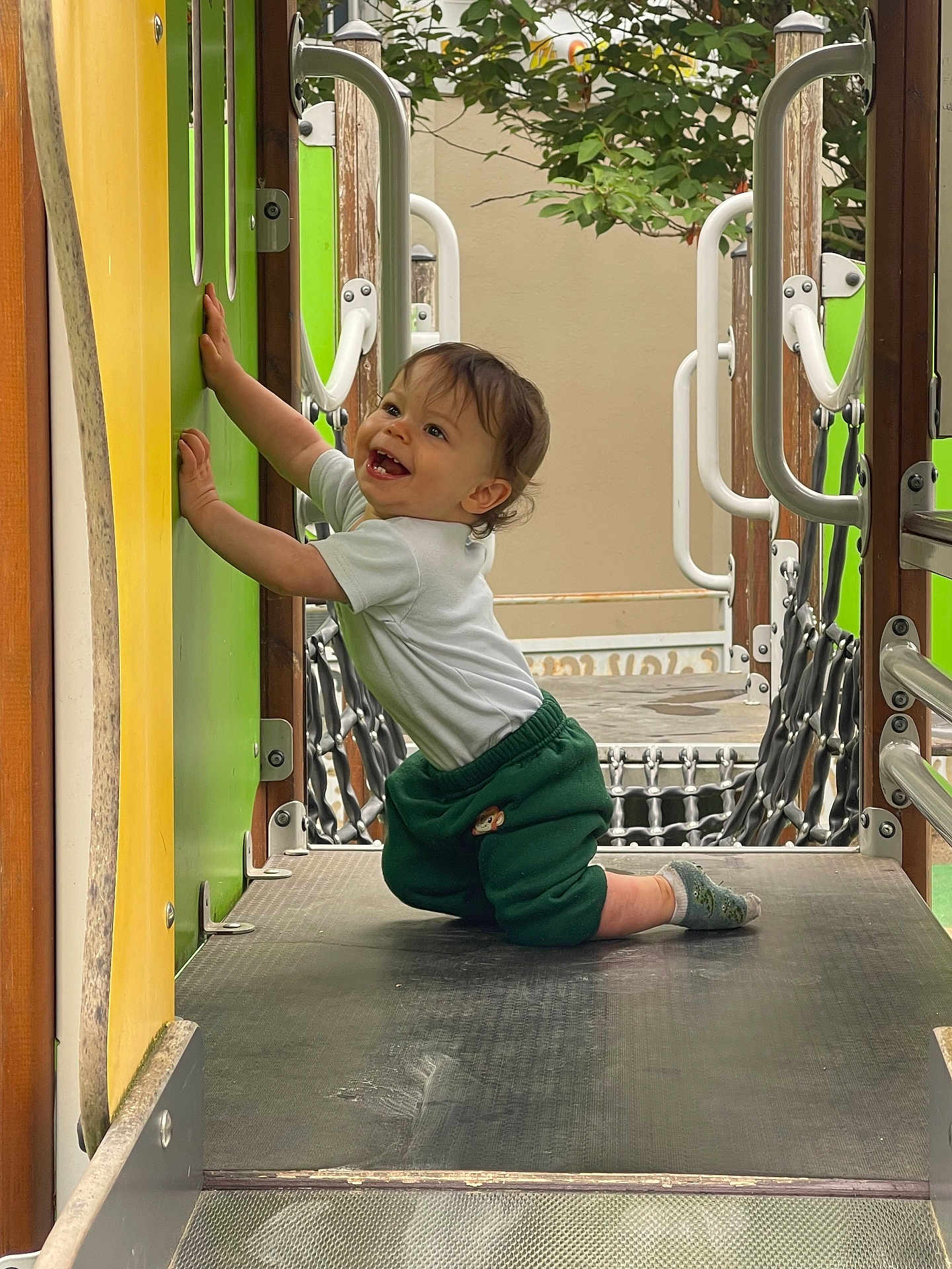 Alessio participe au concours pour gagner de l'argent avec cette photo : toddler, child, playground, smiling, kneeling, green, white_shirt, green_pants, outdoor, play_equipment, metal, wood, handrail, tree, happy, person, sock, platform, crawling, daylight