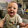 baby, child, smiling, happy, tshirt, sitting, hand_holding, living_room, couch, toys, television, carpet, lamp, bookshelf, fireplace, person, indoor, colorful_text, shorts, home