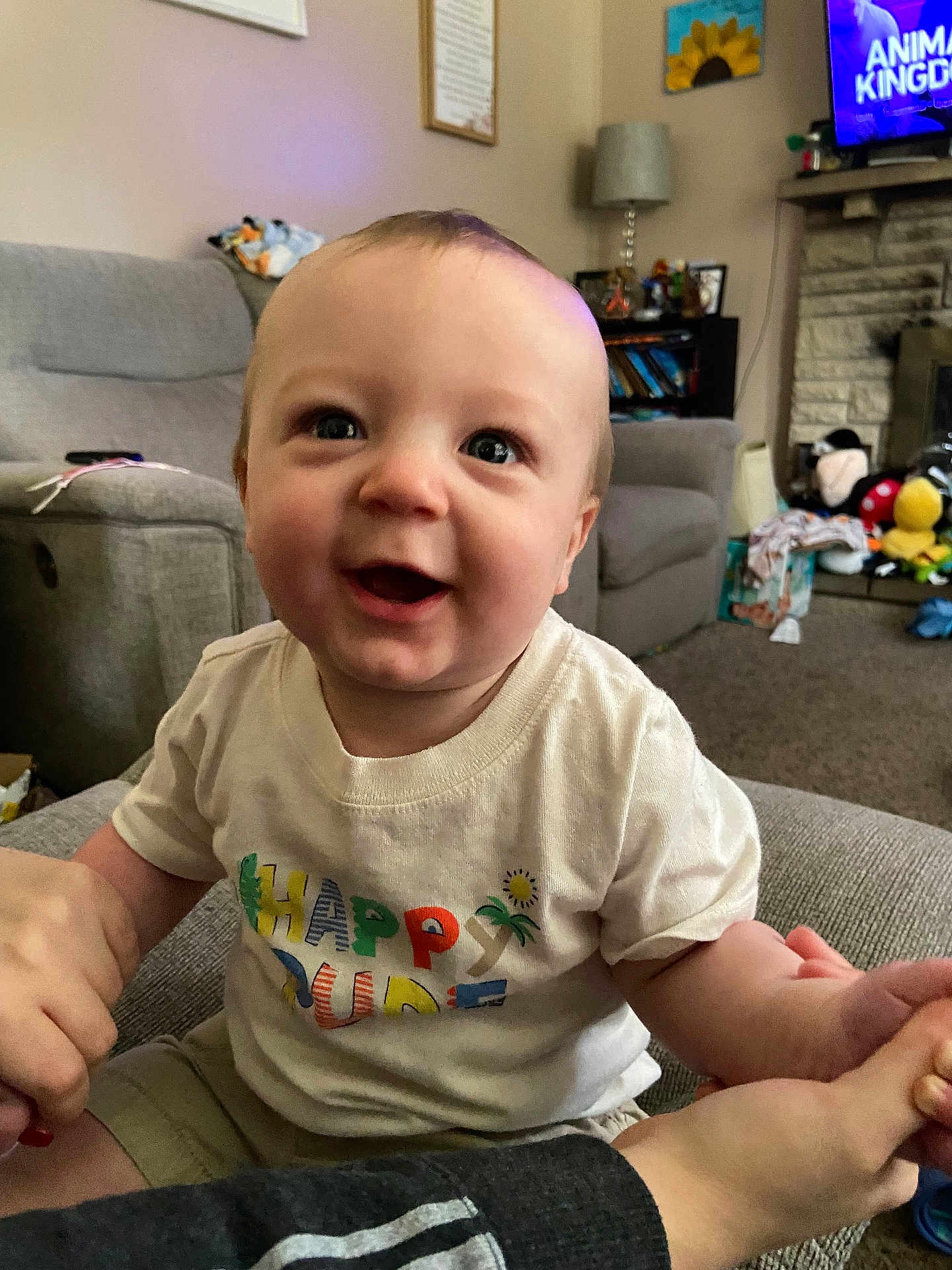 Jensen joined the competition — help win amazing prizes! baby, child, smiling, happy, tshirt, sitting, hand_holding, living_room, couch, toys, television, carpet, lamp, bookshelf, fireplace, person, indoor, colorful_text, shorts, home