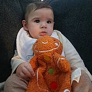Ezekiel is registered to the contest to win money with this photo: baby, child, plush_toy, gingerbread_man, couch, dark_fabric, clothing, bib, striped_pants, hand, face, eyes, soft_toy, indoor, seated, cute, infant, expression, comfort, portrait