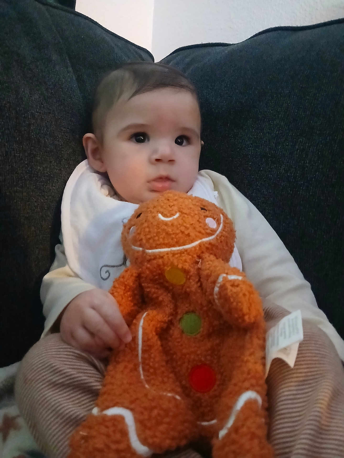 Ezekiel is registered to the contest to win money with this photo: baby, child, plush_toy, gingerbread_man, couch, dark_fabric, clothing, bib, striped_pants, hand, face, eyes, soft_toy, indoor, seated, cute, infant, expression, comfort, portrait