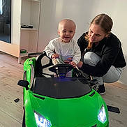 Maylo a rejoint le concours — aidez-le/la à gagner de superbes lots ! baby, child, toy_car, green_car, indoor, wooden_floor, adult, woman, smiling, happy, steering_wheel, crouching, supporting, playtime, home, casual_clothing, light, furniture, joy, interaction