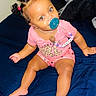 baby, child, pacifier, pink_clothing, hair_buns, bed, indoor, cute, sitting, blue_bedspread, beads, face, person, infant, happy, young, toddler, skin, bright_eyes, cute_hair