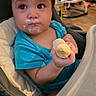 baby, child, toddler, high_chair, banana, food, eating, messy_face, yogurt, blue_shirt, sitting, indoors, home, kitchen, playground_equipment, baby_gear, portrait, close_up, brown_hair, funny