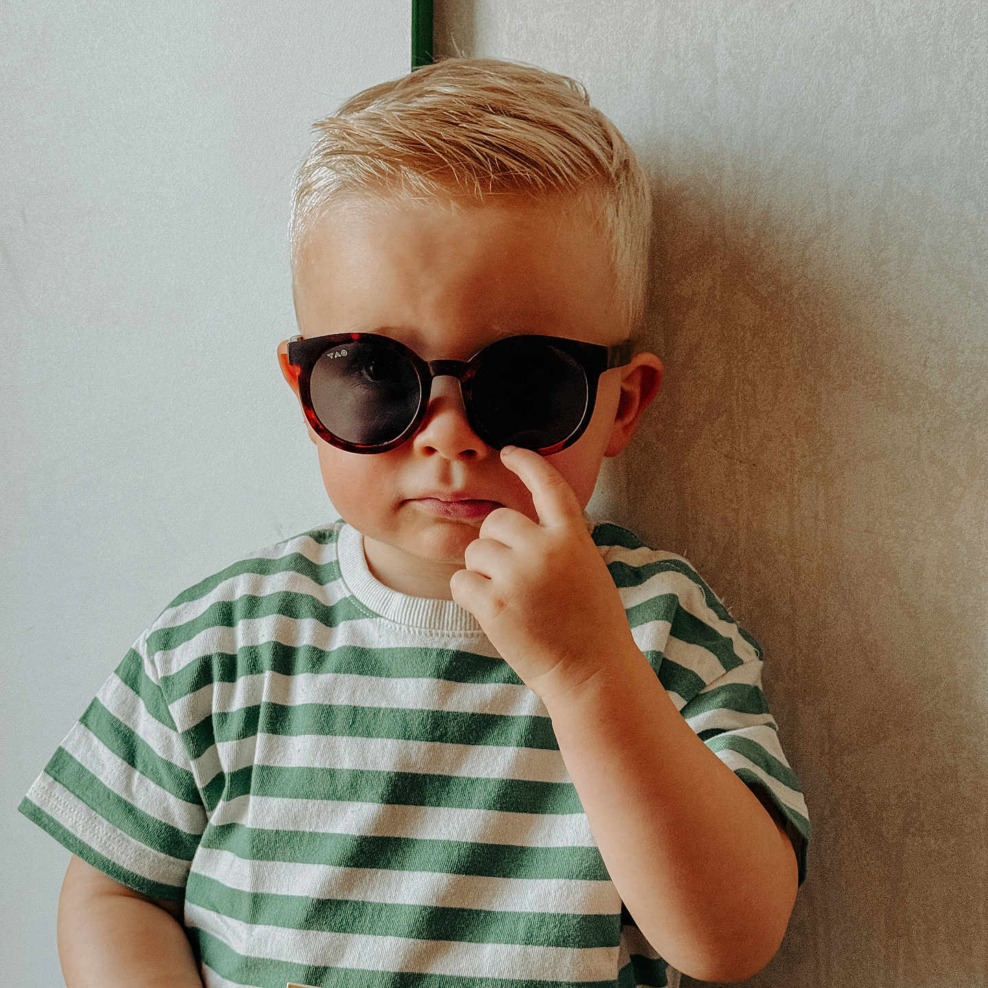 Gautier a rejoint le concours — aidez-le/la à gagner de superbes lots ! blond_hair, casual_clothing, child, fashion, hand, holding_object, indoor, light_skin, mustela, person, portrait, serious_expression, short_hair, standing, striped_shirt, sunglasses, sunscreen, toddler, wall, young_child