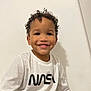 child, toddler, smiling, curly_hair, nasa_shirt, white_background, portrait, happy, cute, sitting, indoors, looking_at_camera, teeth, cheeks, brown_eyes, skin, long_sleeve_shirt, logo, shadow, casual