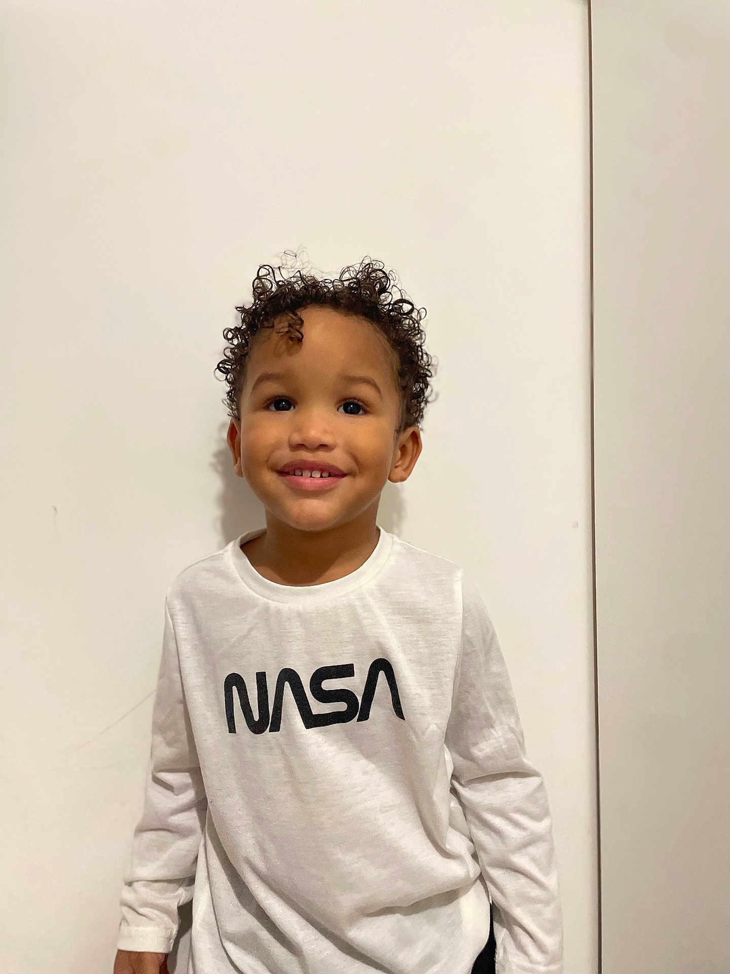 Tidiane a rejoint le concours — aidez-le/la à gagner de superbes lots ! child, smiling, curly_hair, white_wall, nasa_logo, tshirt, portrait, indoors, cute, standing, young_child, happy, teeth, eyes, skin, hair, casual_clothing, minimalist, studio, vertical_photo