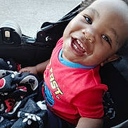 Lavar is registered to the contest to win money with this photo: child, toddler, smile, red_shirt, blanket, stroller, happy, face, curly_hair, outdoor, baby, clothing, person, seat, joy, portrait, cute, hand, patterned_blanket, closeup