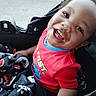 child, toddler, smile, red_shirt, blanket, stroller, happy, face, curly_hair, outdoor, baby, clothing, person, seat, joy, portrait, cute, hand, patterned_blanket, closeup