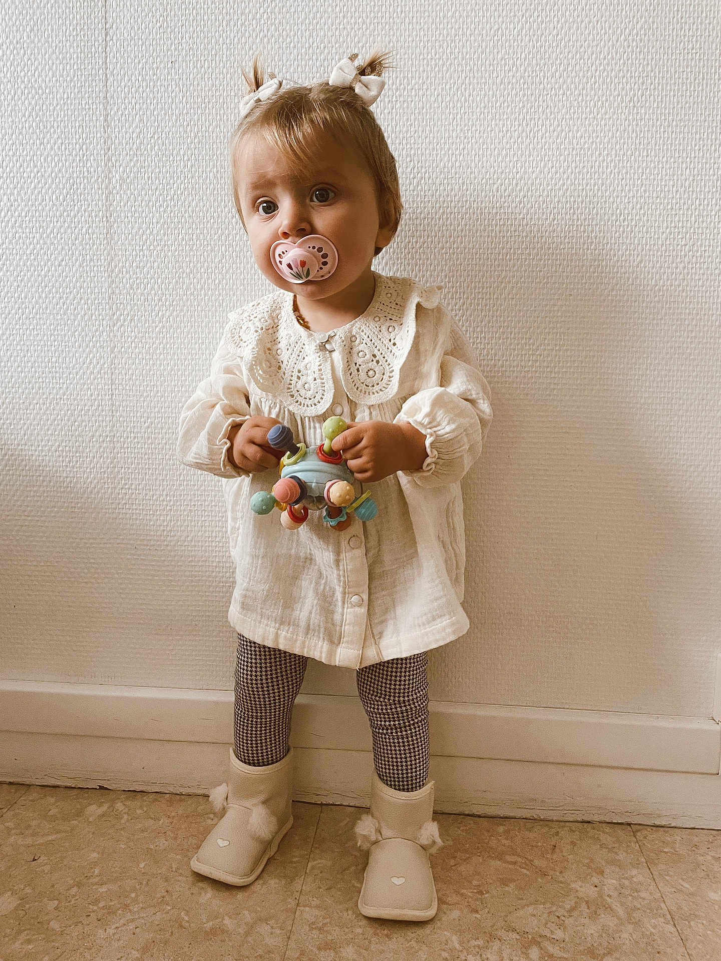 Thaïs a rejoint le concours — aidez-le/la à gagner de superbes lots ! toddler, child, pacifier, toy, white_blouse, lace_collar, pigtails, hair_bows, boots, checkered_leggings, standing, indoors, textured_wall, floor, cute, young_child, portrait, innocence, clothing, person