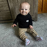 Liam is registered to the contest to win money with this photo: toddler, child, smile, sitting, carpet, black_shirt, beige_pants, white_socks, indoor, wooden_furniture, wood_paneling, cute, baby, floor, person, portrait, happy, home, young_child, clothing