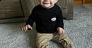 Liam is registered to the contest to win money with this photo: baby, beige_pants, black_shirt, carpet, child, clothing, cute, floor, happy, home, indoor, person, portrait, sitting, smile, toddler, white_socks, wood_paneling, wooden_furniture, young_child