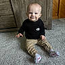toddler, child, smile, sitting, carpet, black_shirt, beige_pants, white_socks, indoor, wooden_furniture, wood_paneling, cute, baby, floor, person, portrait, happy, home, young_child, clothing