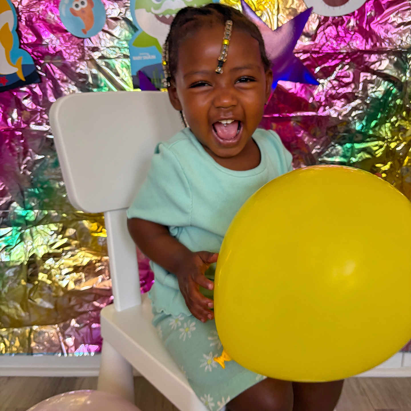 Erza is registered to the contest to win money with this photo: toddler, child, balloon, yellow_balloon, chair, white_chair, smiling, laughing, party, decorations, colorful_background, cartoon_characters, indoors, clothing, socks, shoes, floor, braided_hair, joyful, celebration
