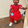 toddler, child, red_dress, beads, hair, smiling, happy, indoor, carpet, sippy_cup, white_shoes, person, standing, banister, handrail, playful, cute, young_child, expression, colorful