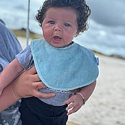 Kaitlyn joined the competition — help win amazing prizes! baby, child, curly_hair, blue_bib, gray_shirt, black_pants, bracelet, hand, adult_hand, outdoor, beach, sand, cloudy_sky, portrait, person, expression, cute, infant, holding, curious