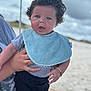 baby, child, curly_hair, blue_bib, gray_shirt, black_pants, bracelet, hand, adult_hand, outdoor, beach, sand, cloudy_sky, portrait, person, expression, cute, infant, holding, curious