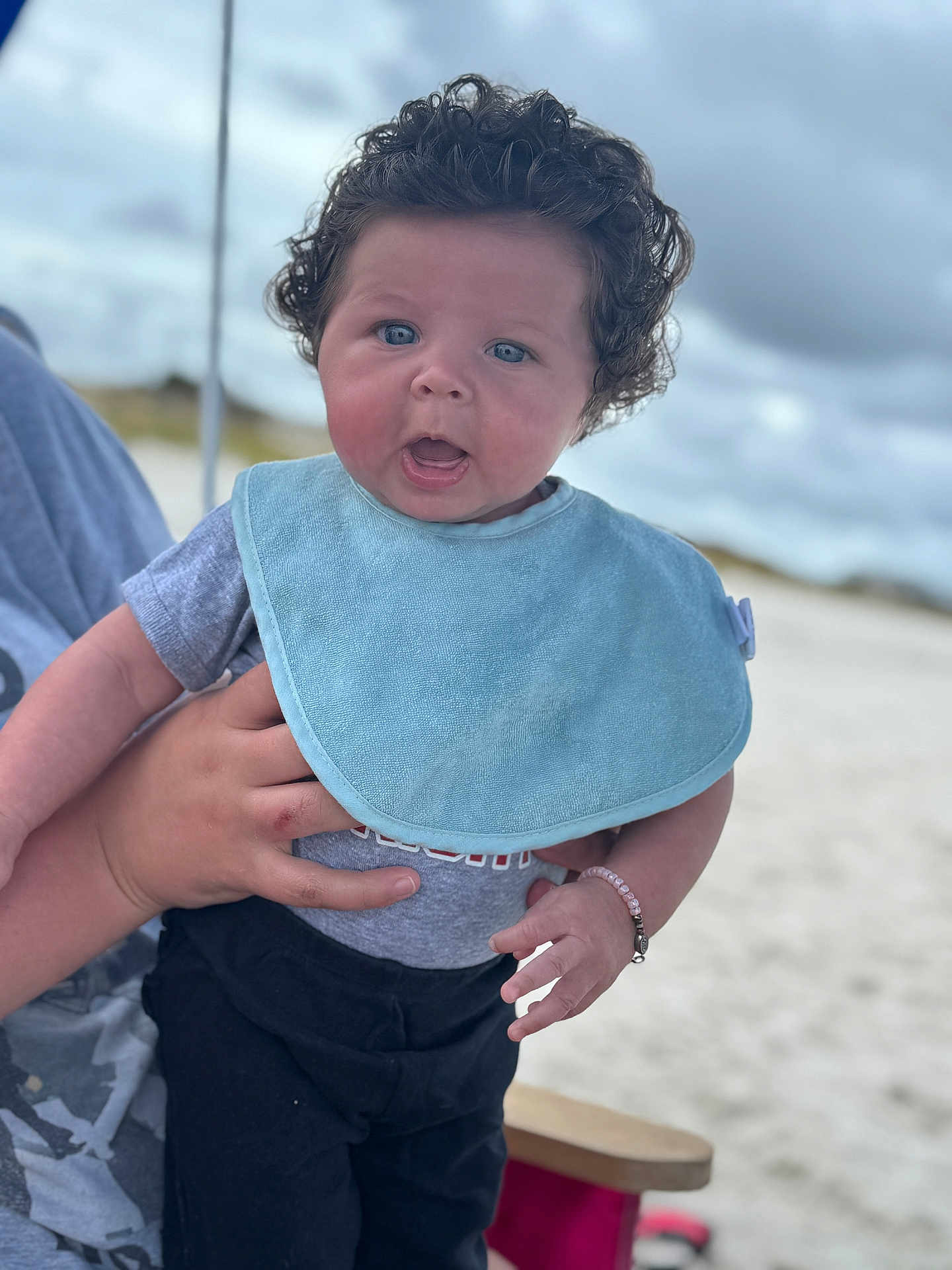 Kaitlyn joined the competition — help win amazing prizes! baby, child, curly_hair, blue_bib, gray_shirt, black_pants, bracelet, hand, adult_hand, outdoor, beach, sand, cloudy_sky, portrait, person, expression, cute, infant, holding, curious