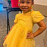 child, girl, yellow_dress, smile, indoor, carpet, wall, table, red_bin, hair_beads, puffs, posing, happy, fashion, cute, person, standing, room, colorful, portrait