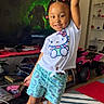 child, smiling, hello_kitty, shorts, tshirt, indoor, colorful, hair_accessories, toy_car, rug, television, furniture, playful, cute, white_shoes, pink_socks, cabinet, room, happy, standing