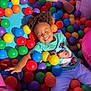 child, ball_pit, colorful_balls, play, happy, smiling, curly_hair, casual_clothing, indoor_play_area, fun, leisure, bright_colors, pink_cushions, blue_socks, purple_balls, green_balls, yellow_balls, orange_balls, red_balls, two_children