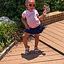child, girl, sunglasses, heart_shaped, pink_shirt, navy_skirt, popsicle, summer, outdoor, deck, wooden_floor, plants, greenery, brick_wall, sunlight, shadows, sandals, cute, posing, happy
