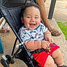 Ezekiel is registered to the contest to win money with this photo: baby, stroller, smiling, child, outdoor, concrete, grass, person, legs, footwear, leopard_print, keychain, red_shorts, patterned_shirt, black_stroller, happy, cute, sitting, casual, daytime