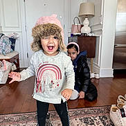 Aarya is registered to the contest to win money with this photo: toddler, child, hat, pink_hat, sweater, rainbow, black_coat, headband, indoor, carpet, wood_floor, lamp, bag, shoes, door, expression, happy, grumpy, person, home