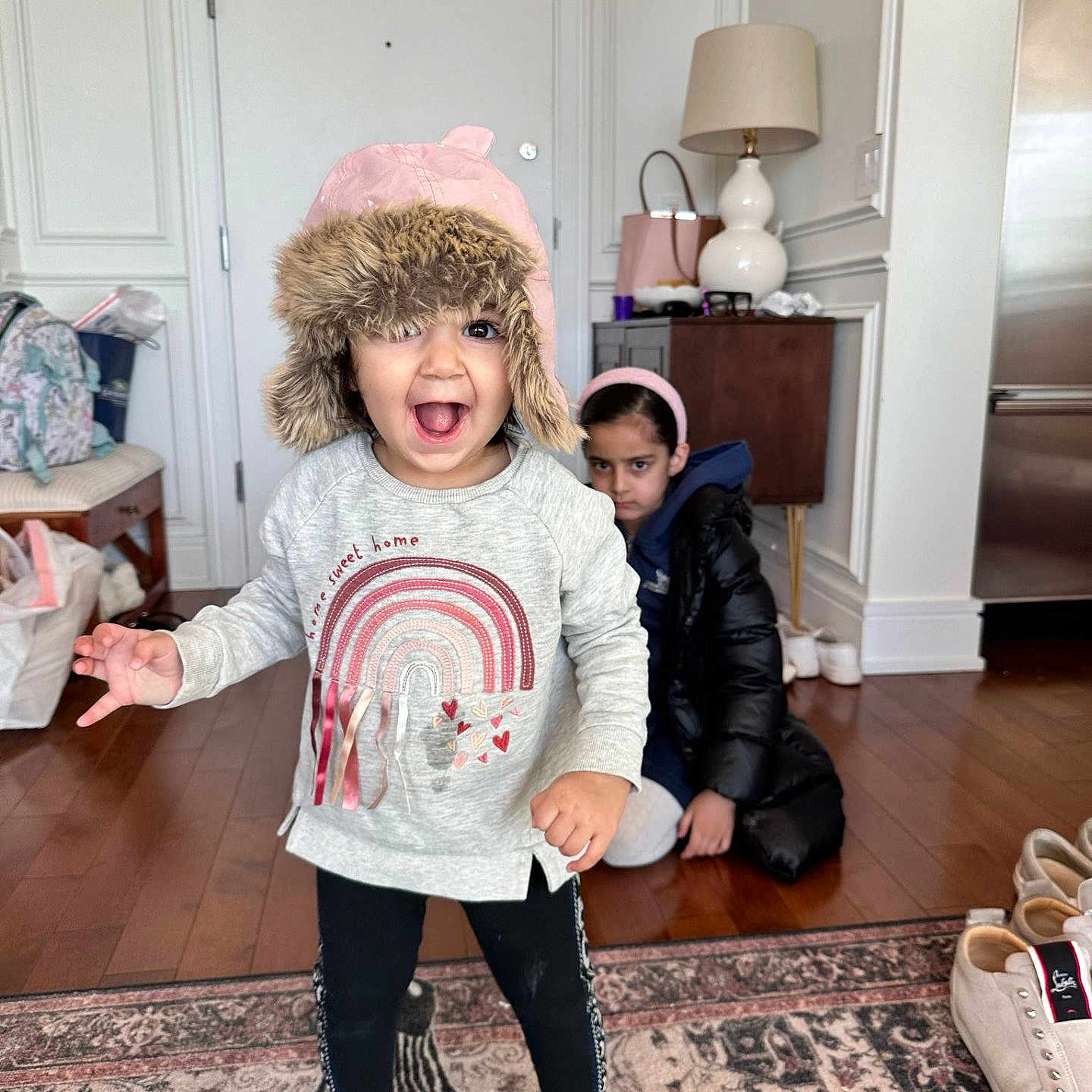 Aarya is registered to the contest to win money with this photo: toddler, child, hat, pink_hat, sweater, rainbow, black_coat, headband, indoor, carpet, wood_floor, lamp, bag, shoes, door, expression, happy, grumpy, person, home