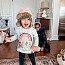 toddler, child, indoor, hat, sweater, rainbow, floor_rug, expression, smile, serious, lamp, table, shoes, headband, black_jacket, wood_floor, door, bag, person, home_interior