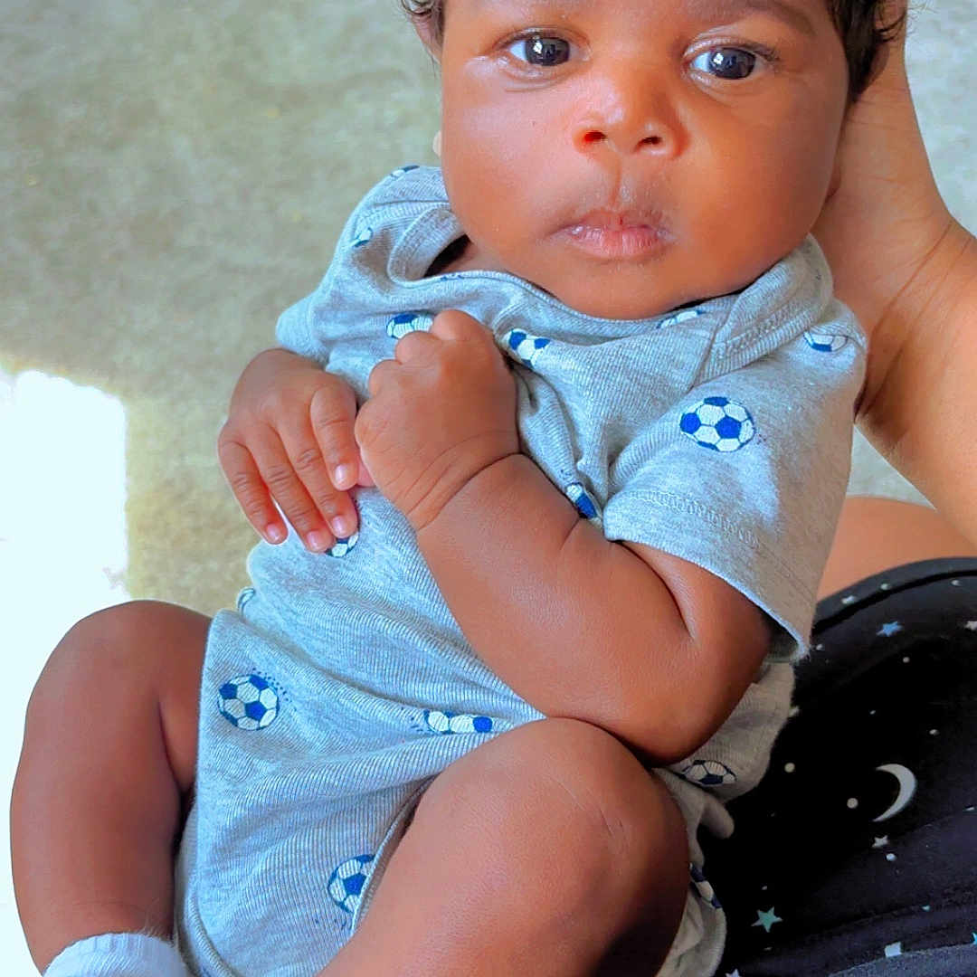 Tyaire is registered to the contest to win money with this photo: baby, infant, child, onesie, socks, hand, face, curly_hair, indoor, person, cute, holding, looking, eyes, skin, legs, clothing, pattern, background, soft_lighting
